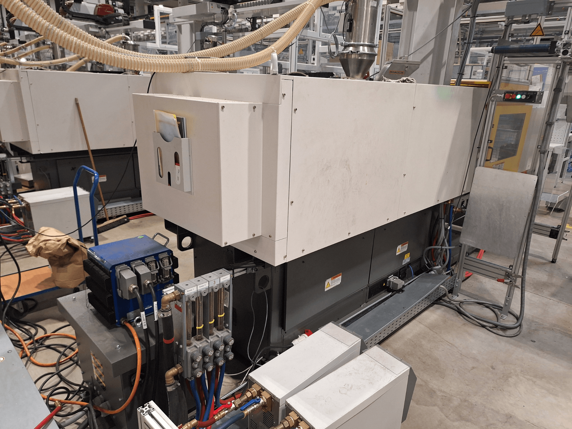 Apparecchiature industriali in un ambiente di fabbrica; comprende una grande macchina con pannello di controllo, tubi e collegamenti elettrici, vista laterale.