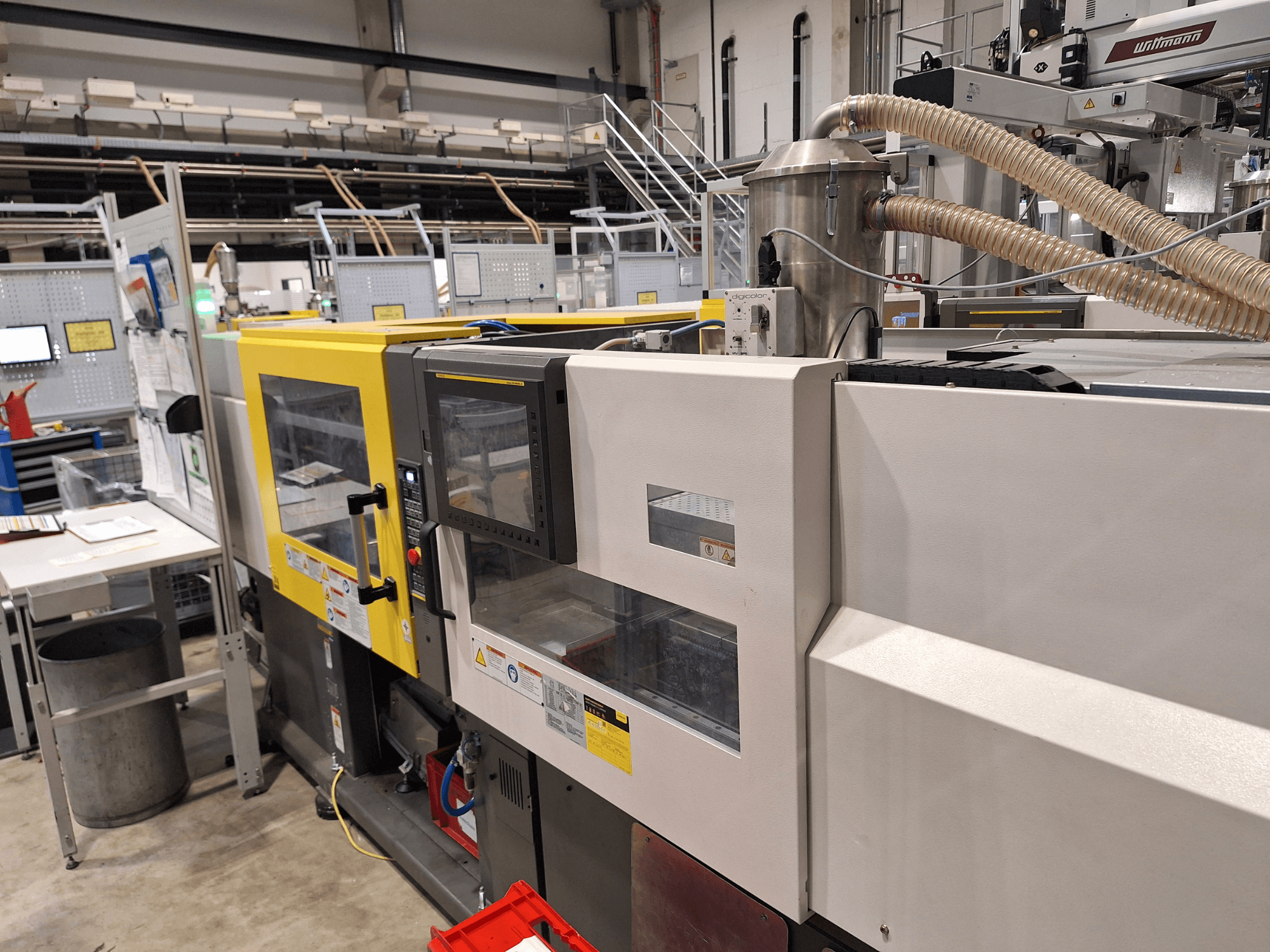 Vista frontale di una pressa a iniezione multicolore, con sezioni gialle e grigie, con pannello di controllo e sistema di scarico.