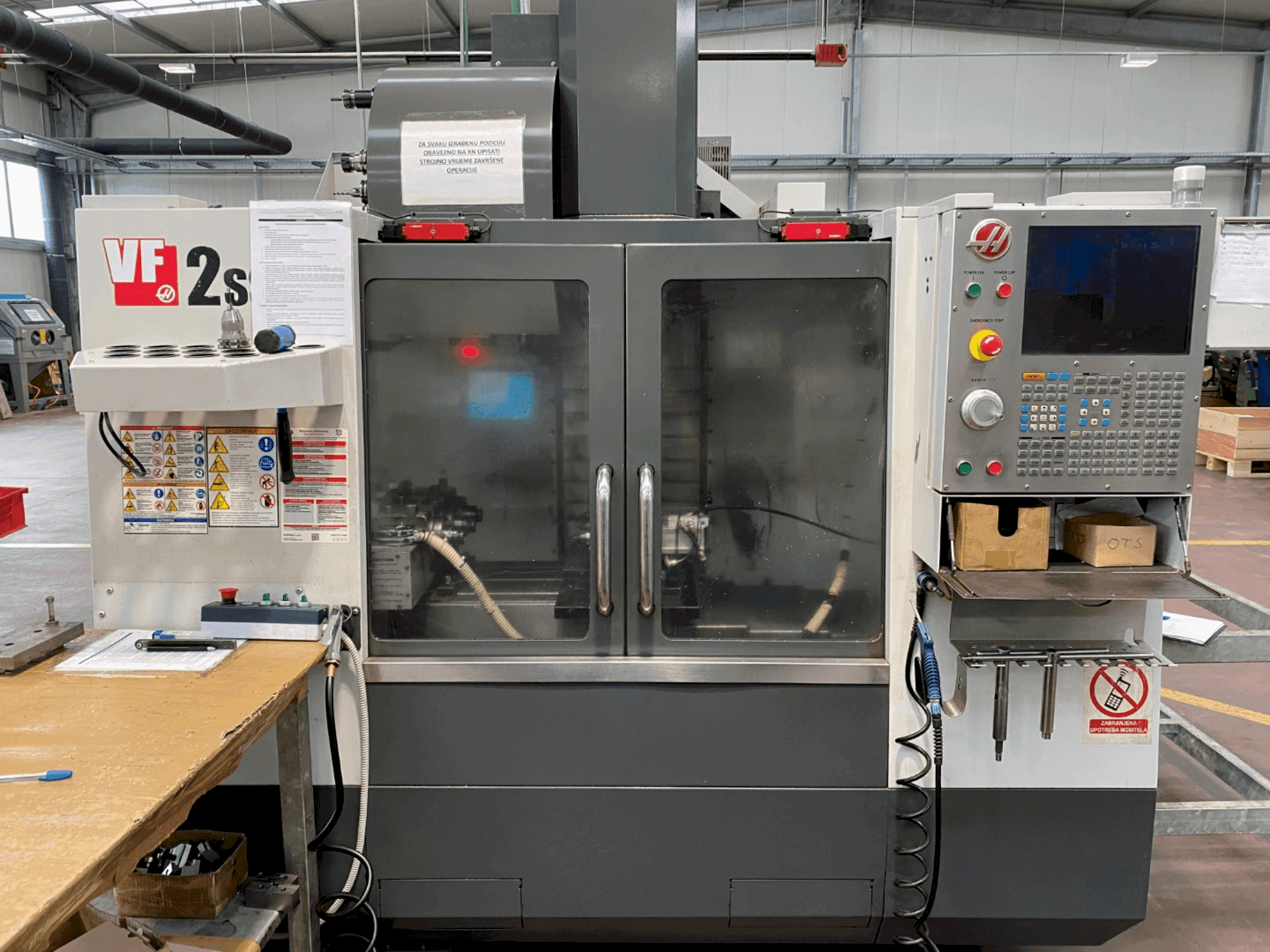 Centro di lavoro Haas VF-2SS in vista frontale, con pannello di controllo, tubo del refrigerante ed etichette di sicurezza sul lato.