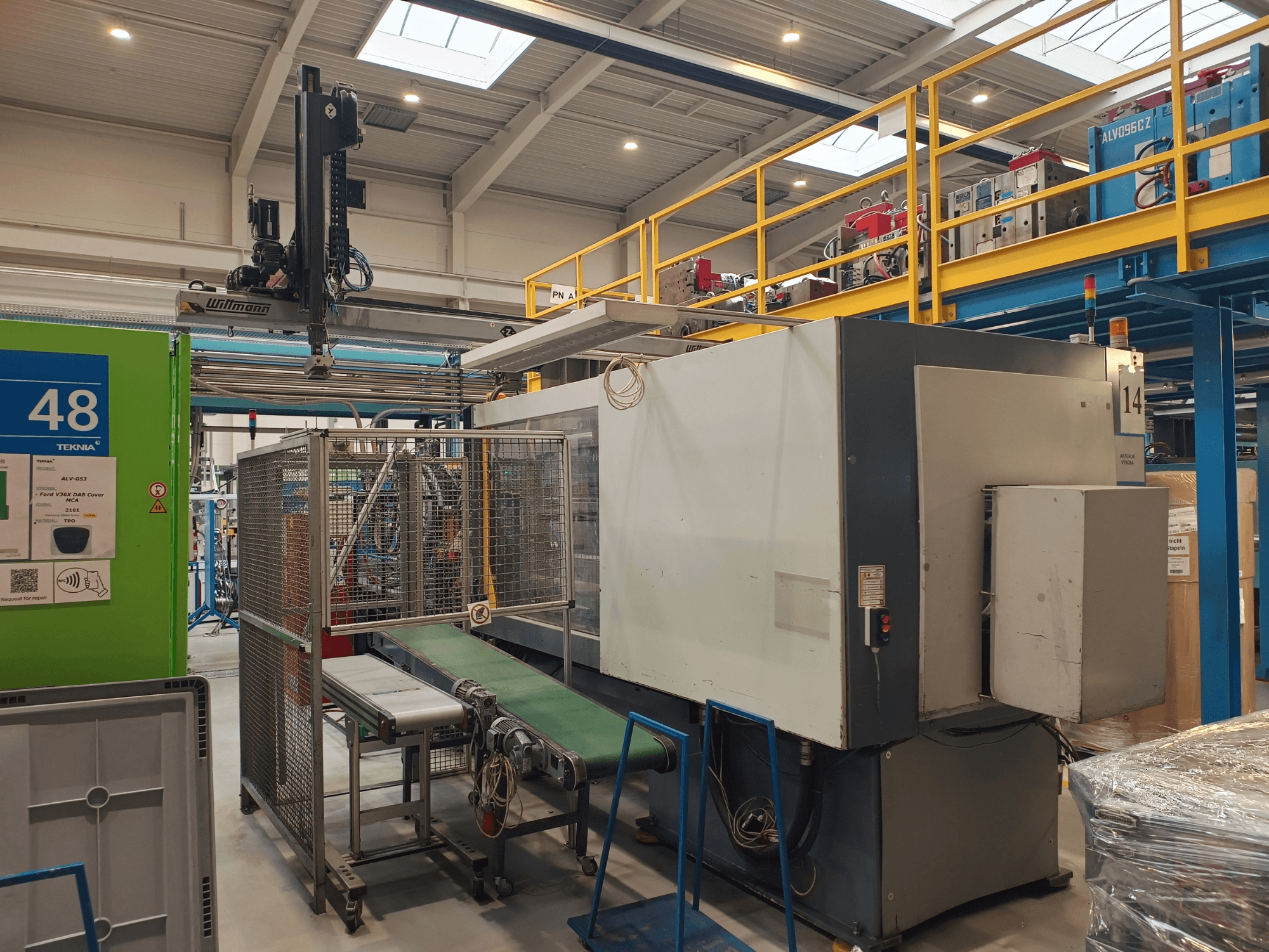 Apparecchiatura di automazione industriale con trasportatore verde, etichettato "48", e carroponte; vista laterale della macchina sul pavimento della fabbrica.